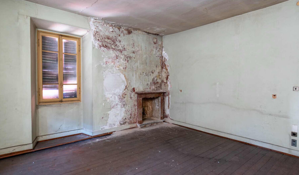 Salone al primo piano di Casa Varenna a Locarno prima del restauro, con camino storico in pietra, pavimento in assi di legno e intonaci deteriorati.