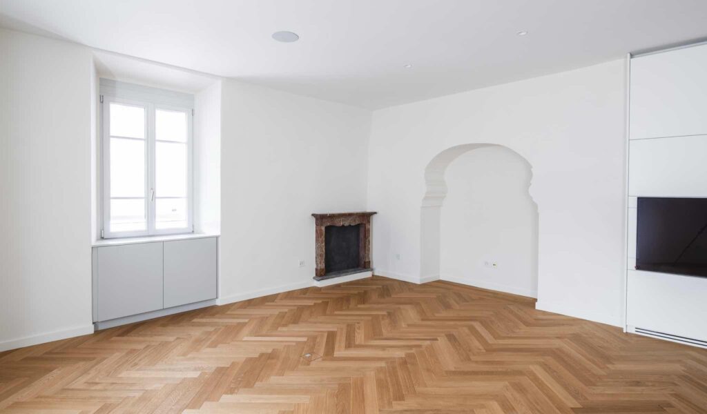 Salone al primo piano di Casa Varenna a Locarno dopo il restauro, con camino storico in pietra, nicchia ad arco e pavimento in parquet a spina di pesce.