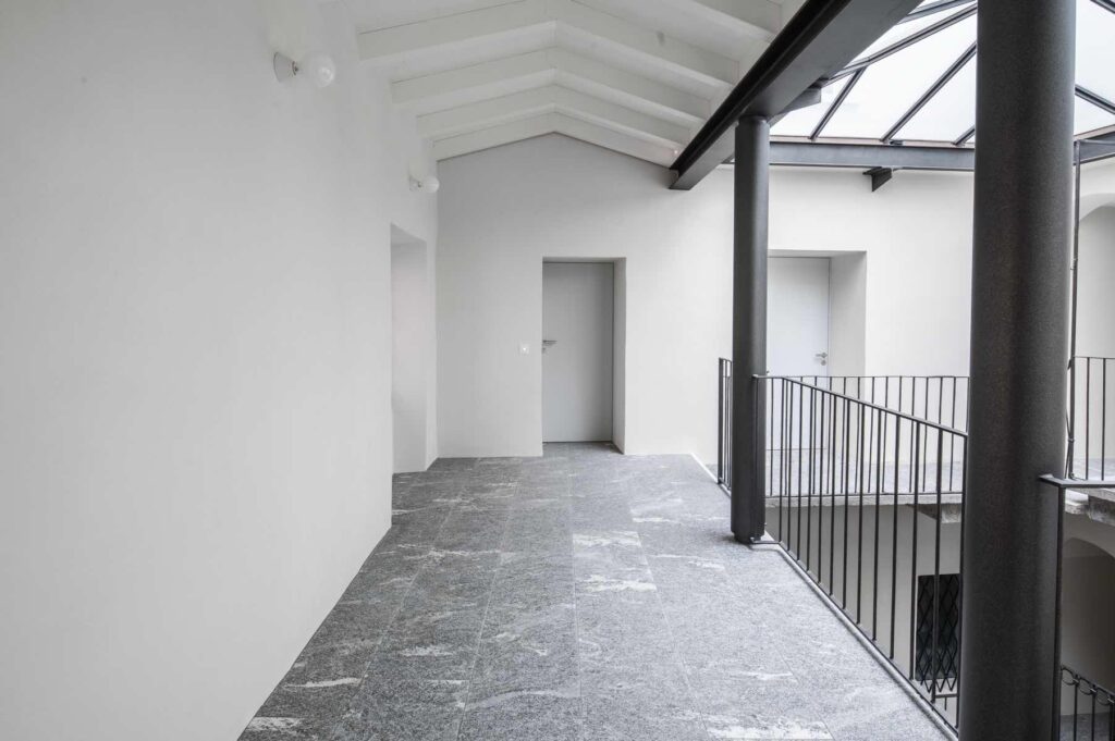 Ballatoio al secondo piano di Casa Varenna a Locarno dopo il restauro, con pavimentazione in pietra, parapetti metallici e copertura vetrata della corte interna.