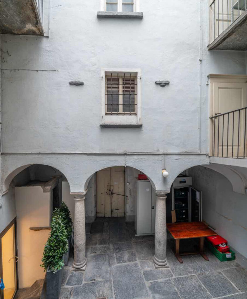 Corte interna di Casa Varenna a Locarno prima del restauro, con porticato ad archi, colonne in pietra, pavimentazione in lastre di pietra e ballatoi.
