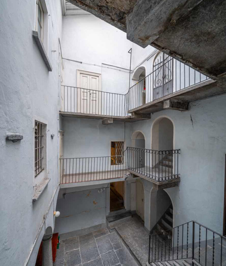 Corte interna di Casa Varenna a Locarno prima del restauro, con ballatoi su più livelli, ringhiere in ferro, archi e pavimentazione in pietra.