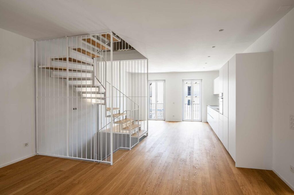 Interno della maisonette di Casa Varenna a Locarno dopo la ristrutturazione, con scala a gradini in legno, parapetti metallici bianchi e cucina contemporanea.